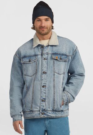Man som bär en blå stickad mössa, en ljusblå denimjacka med sherpa-krage och blå byxor, med ena handen i jackfickan, mot en enfärgad bakgrund.