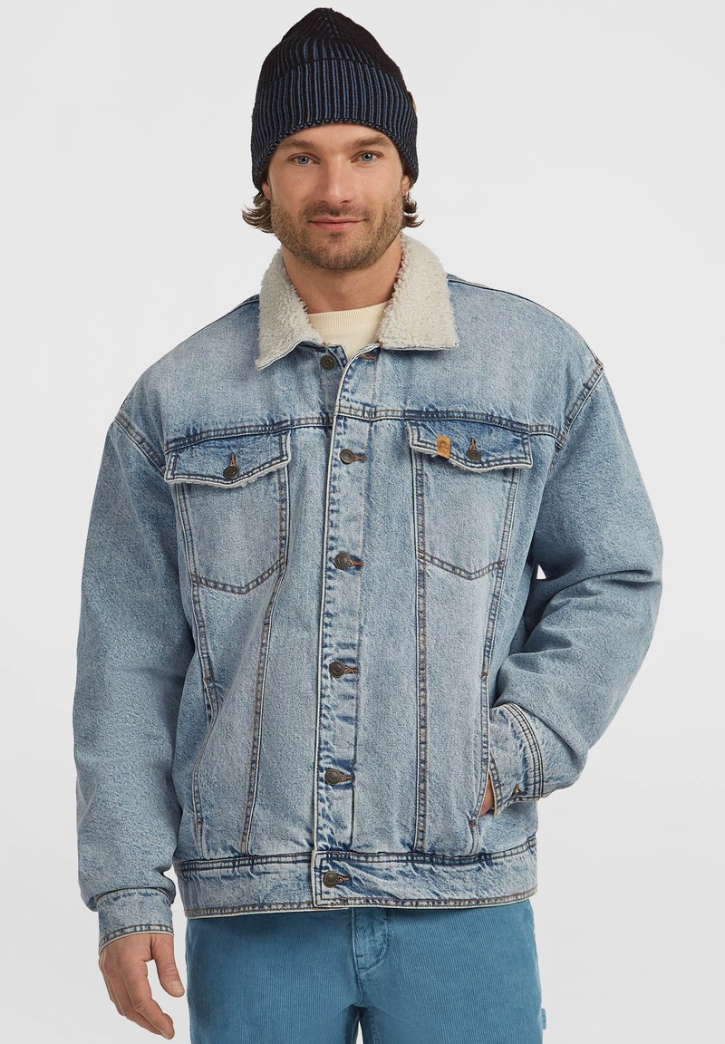 Homme portant un bonnet en tricot bleu, une veste en denim avec col en sherpa bleu clair et un pantalon bleu, avec une main dans la poche de la veste, sur fond uni.