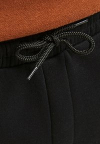 Pantaloni jogger neri con elastico in vita e cordoncino nero, caratterizzati da una superficie testurizzata; sopra c'è una sezione di maglione arancione.