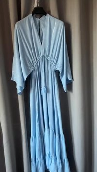 Ljusblå klänning med djup V-ringning, breda ärmar, sammetsliknande midja och lager-på-lager fåll. Tillverkad av mjuk, flödande tyg med subtil lyster.