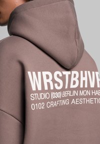 Kapuzensweatshirt in Taupe mit weißem Druck auf dem Rücken. Verfügt über einen weichen, strukturierten Stoff und einen lässigen Schnitt.