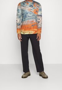 Mehrfarbiges Langarmhemd mit abstrakten Spiralen in Blau, Orange und Creme; kombiniert mit navyblauen Hosen und braunen Schuhen.