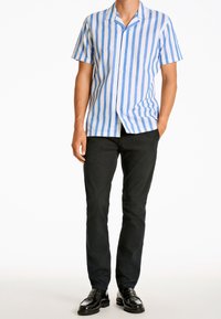 Koszula na krótki rękaw na guziki w białym kolorze z niebieskimi pionowymi paskami, zestawiona z czarnymi spodniami i błyszczącymi czarnymi butami. Czysty, casualowy wygląd.