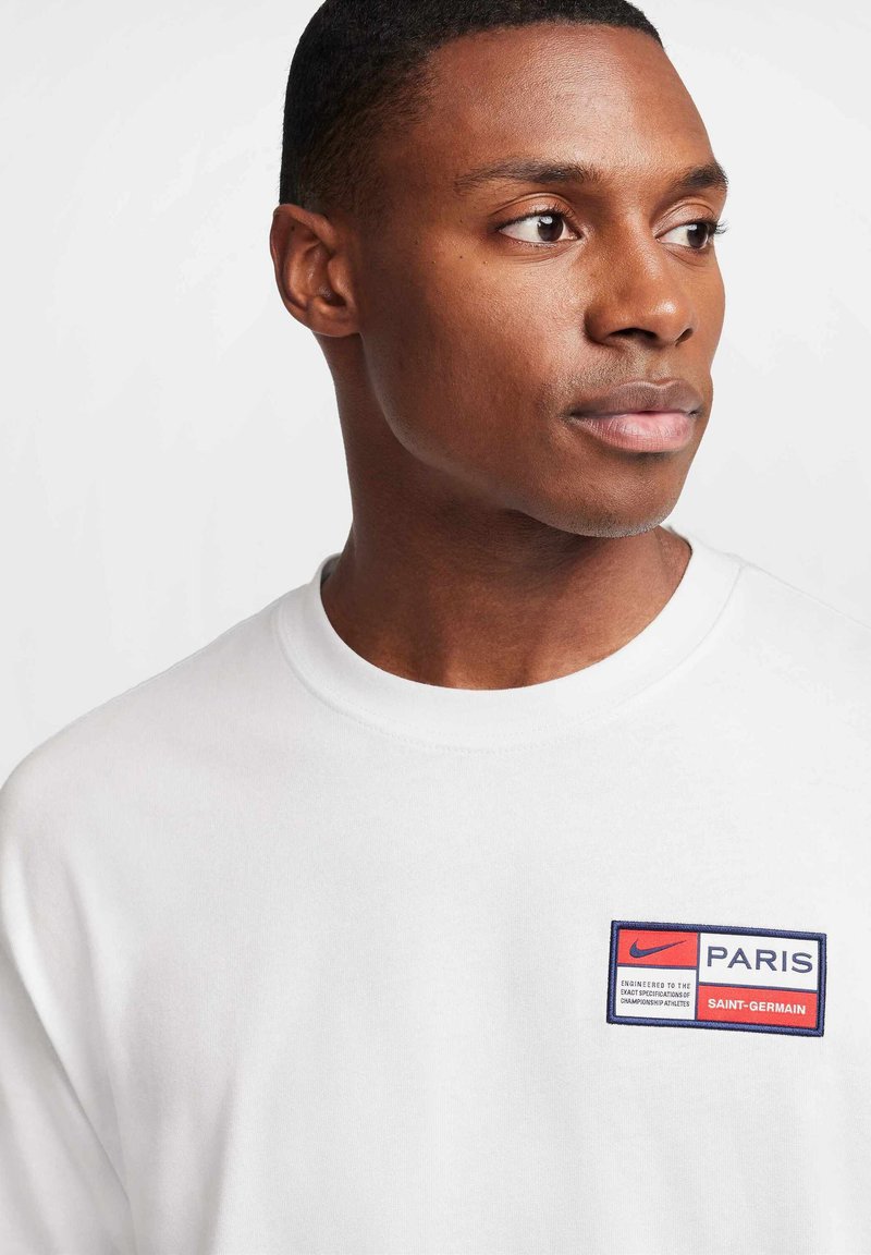 Biała bawełniana bluza z okrągłym dekoltem. Posiada czerwoną i niebieską naszywkę na piersi z napisem "Nike Paris Saint-Germain". Gładka tekstura.