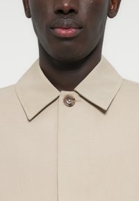 Μπεζ παλτό με δομημένο, φαρδύ γιακά, κούμπωμα με ένα κουμπί και λεία υφή υφάσματος, με ορατές ραφές στο μπροστινό μέρος.