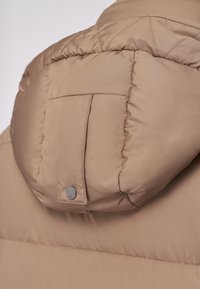 Giubbotto imbottito in beige chiaro con una texture trapuntata, dotato di cappuccio arrotondato e dettaglio con bottone a pressione sulla tasca posteriore.