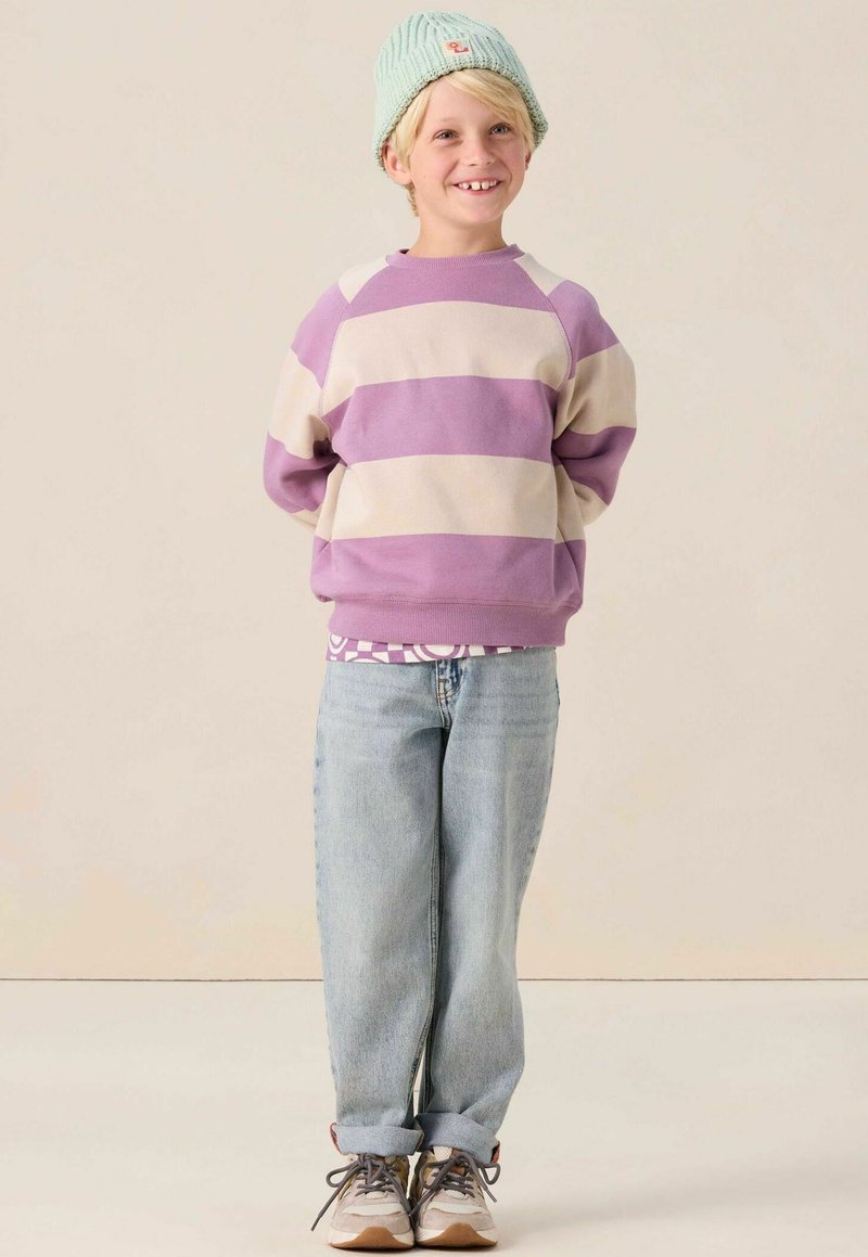 Sweat-shirt rayé violet et crème, jean bleu clair et baskets grises. Enfant portant un bonnet en tricot vert clair. Souriante et debout.