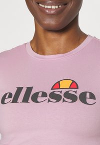 Světle růžové tričko s kulatým výstřihem a velkým černým logem "ellesse", s barevným slunečním emblémem nad textem. Hladká textura.