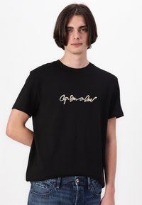 Camiseta de algodón negra de manga corta, con texto en cursiva color crema en la parte frontal. Diseño clásico de cuello redondo.