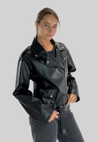 Veste en cuir synthétique noir avec manches surdimensionnées, boutons-pression argentés, deux poches avant et taille ceinturée. Texture lisse et finition brillante.