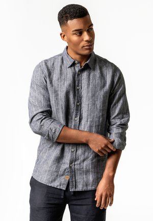 Junger Mann, der den aufgerollten Ärmel seines grauen, gestreiften Hemds mit Knöpfen anpasst, trägt dunkle Hosen und steht vor weißem Hintergrund.