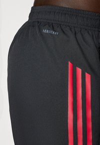 Shorts de sport noirs avec une texture lisse, présentant trois bandes verticales rouges sur le côté et un logo "AEROREADY" près de la taille.