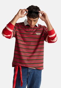 Camiseta de manga larga a rayas en rojo, verde y negro, con cuello blanco y logo, con un detalle rojo atado en el dobladillo.