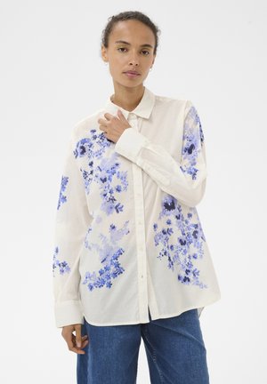 Woman wearing loose white button-up shirt with blue floral patterns and blue jeans, standing against a plain light background.