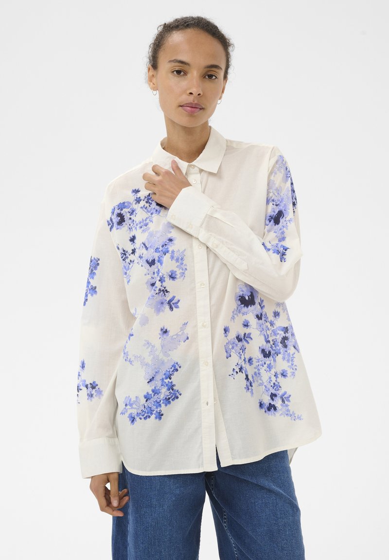 Femme portant une chemise blanche ample à boutons avec des motifs floraux bleus et un jean bleu, debout devant un fond clair uni.