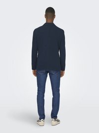 Marinblå kavaj med skräddarsydd passform, i kombination med blå jeans och vita sneakers, sedd från baksidan som framhäver axel- och kavajdesign.