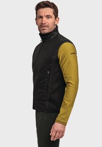 Schwarwestes gelbes Thermoweste mit einer Reißverschlusstasche, ausgestattet mit einer gesteppten Oberseite und gerippten unteren Ärmel, getragen über einem Langarmshirt.