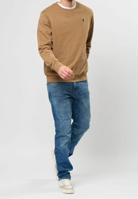 Sudadera de cuello redondo marrón hecha de algodón, combinada con pantalones vaqueros desteñidos azules. Zapatillas blancas complementan el atuendo casual. Diseño sencillo y limpio.