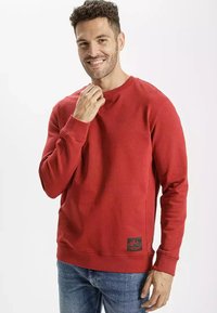 Czerwony sweter z okrągłym dekoltem, wykonany z miękkiego materiału, z prążkowanymi mankietami i dołem, z czarną łatą z logo na dolnej lewej stronie.