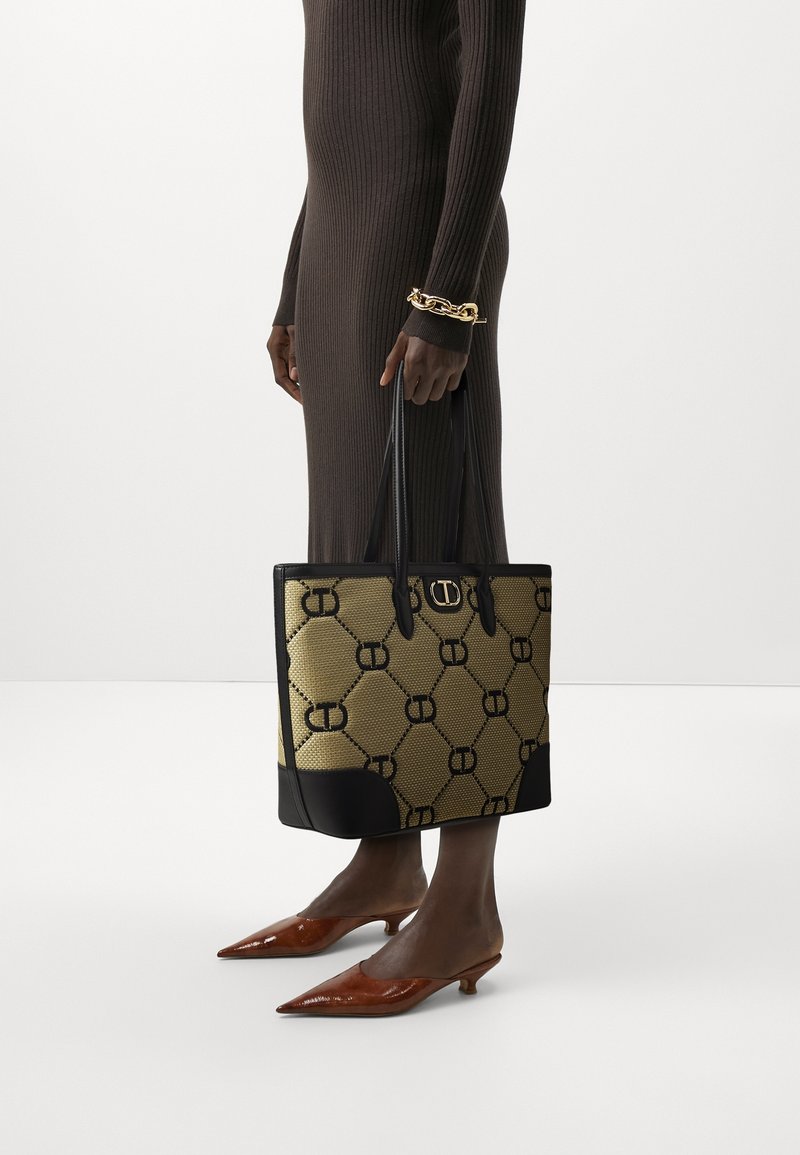 Sac en toile avec un motif géométrique noir et or, base en cuir noir, et doubles longues poignées. Porté avec une robe marron ajustée.