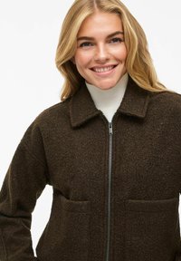 Veste zippée marron en tissu texturé, avec un col et deux poches avant. Dotée d'une fermeture éclair foncée et d'une forme structurée.