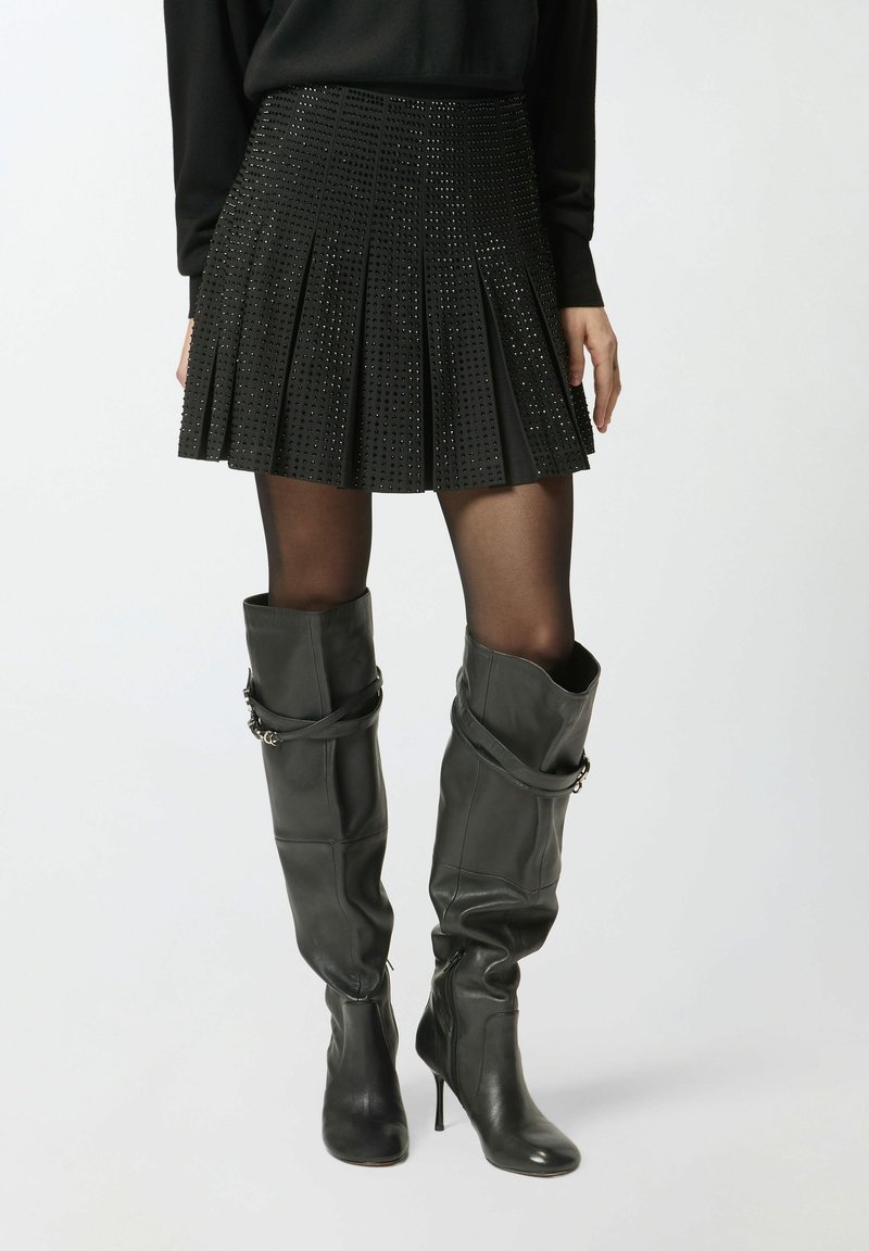 Mini gonna nera plissettata con design borchiato, abbinata a stivali in pelle grigi sopra il ginocchio con tacco alto e dettaglio con cinturino.