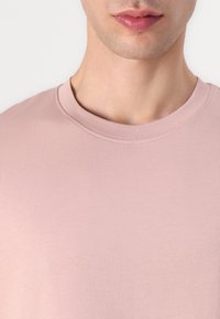 Hellrosa T-Shirt mit Rundhalsausschnitt, glatter, gerippter Textur und kurzen Ärmeln. Nahaufnahme zeigt die Nähte am Ausschnitt.