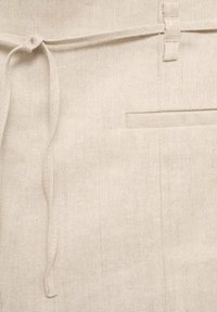 Lniany fartuch w jasnobeżowym kolorze z wiązanym w pasie paskiem, posiadający jedną kieszeń i delikatną teksturę tkaniny.