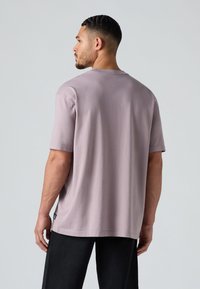 T-shirt à manches courtes, col rond en coton lavande clair. Présente une coupe décontractée avec un petit détail d'étiquette sur le côté. Porté avec un pantalon sombre.
