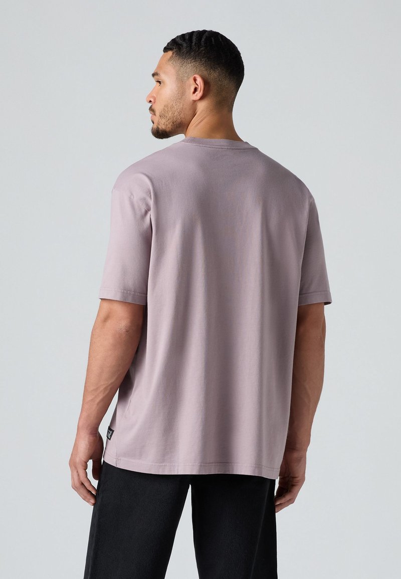 Kurzärmliges T-Shirt mit Rundhalsausschnitt aus leichtem Lavendelbaumwollstoff. Verfügt über eine lockere Passform und ein kleines seitliches Etikett. Kombiniert mit dunklen Hosen.