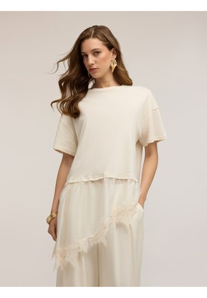 Vrouw met golvend haar die een oversized crème T-shirt draagt over een rok of jurk met kanten rand, gouden blad oorbellen en armbanden op een lichte achtergrond.