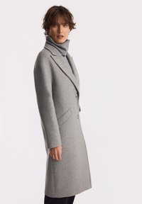 Kobieta z krótkimi kręconymi włosami, ubrana w szary golf i długi szary płaszcz zapinany na guziki, stojąca na tle jednolitego jasnego tła.