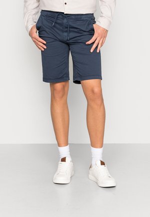 Homme portant un short bleu marine mi-genoux, des chaussettes blanches, des baskets blanches et une chemise à manches longues de couleur claire, debout devant un fond uni.