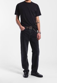 Camiseta negra combinada con jeans de mezclilla negros, que cuentan con un cinturón de hebilla plateada y un acento de cadena. Modelo vistiendo zapatos negros.
