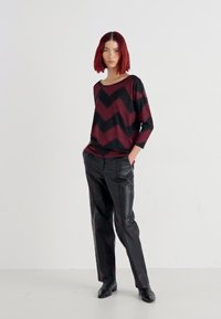 Bordeauxroter und schwarzer, übergroßer Pullover mit Chevron-Muster, kombiniert mit schwarzen Lederhosen. Das Model steht mit einer Hand in der Tasche und trägt schwarze Schuhe.