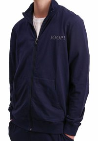 Giacca zip-up blu navy con colletto alto e due tasche frontali. Presenta la scritta "JOOP!" in grigio chiaro sulla parte superiore sinistra. Il materiale sembra liscio.