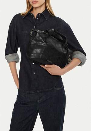 Donna in un completo di denim scuro che tiene una borsa a mano in pelle nera con motivo cucito e logo dorato vicino al centro.