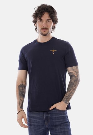 Uomo con capelli ricci che indossa una t-shirt blu navy con un piccolo emblema dorato, mostra tatuaggi sul braccio, posa con una mano in tasca.