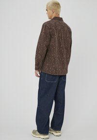 Chemise marron à motif avec une coupe décontractée, dotée d'un col et d'une patte de boutonnage, associée à un jean en denim bleu foncé ample et des baskets beiges.