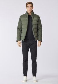 Giacca a vento verde con colletto alto, design trapuntato, abbinata a una maglietta nera a maniche lunghe, jeans neri e sneakers bianche.