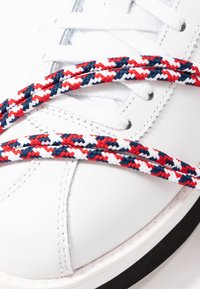 Baskets en cuir blanc avec des lacets tressés rouges, blancs et bleus croisant la zone des orteils contre un bord de semelle noir.