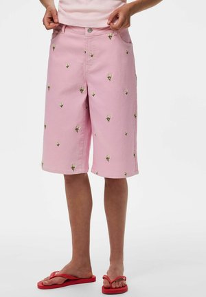 Persoon die roze knielange shorts draagt met kleine witte bloemenprints en rode teenslippers, staand tegen een witte achtergrond.
