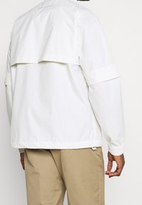 Mężczyzna w białej długo-sieszanej kurtce z tylnym rozcięciem, ubrany w beżowe spodnie, widoczny od tyłu na jednolitym tle.