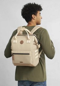 Zaino beige con spallacci imbottiti, tasca anteriore con zip, patch in pelle con logo e dettagli a righe orizzontali sui lati. Texture liscia.