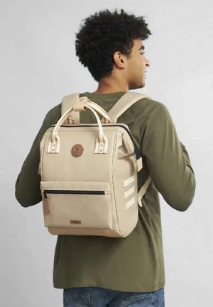 Sac à dos beige avec des bretelles rembourrées, une poche zippée à l'avant, un patch en cuir avec logo et des accents de rayures horizontales sur le côté. Texture lisse.
