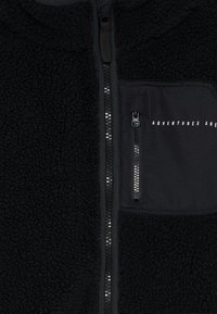 Chaqueta de forro polar negra con cremallera central y bolsillo en el pecho con cremallera, que presenta el texto "AVENTURAS POR DELANTE".