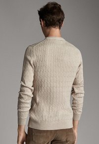 Beige neulottu villapaita, jossa on teksturoitu kaapelikuvio, pitkät hihat ja pyöreä kaula-aukko. Kangas näyttää pehmeältä ja istuvuudeltaan vartalonmyötäiseltä.