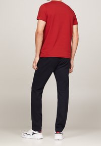 Rød t-shirt med korte ærmer, der er parret med mørkeblå bukser. Modellen har hvide sneakers på med sorte og røde detaljer. Glat stoftekstur.