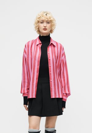 Person med krøllete blondt hår iført en rosa og rødt stripete skjorte med knapper over en svart turtleneck og svart skjørt mot en ensfarget bakgrunn.