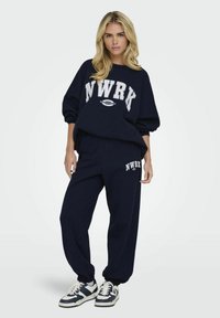 Felpa a girocollo blu navy e pantaloni jogging coordinati con il logo bianco "NWRK", polsini a coste e vestibilità rilassata. Abbinati a sneakers.